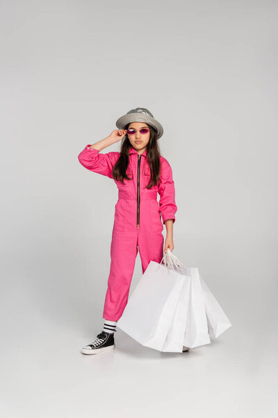 stylish girl in pink outfit and panama hat holding shopping bags on grey, adjusting sunglasses