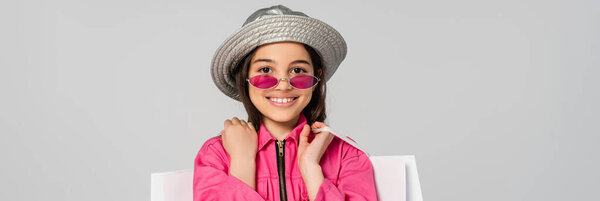 positive girl in stylish outfit, sunglasses and panama hat posing with shopping bags on grey, banner