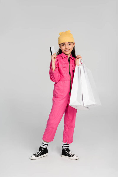 счастливая девушка в шляпе и трикотажном наряде с кредитной картой на сером, детский шопинг, мода, радость