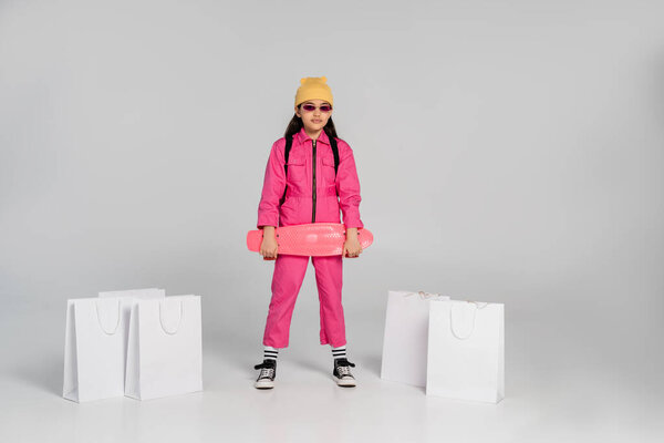 стильная девушка в шапочках и солнцезащитных очках держа доску с пенни рядом с сумками для покупок на сером фоне