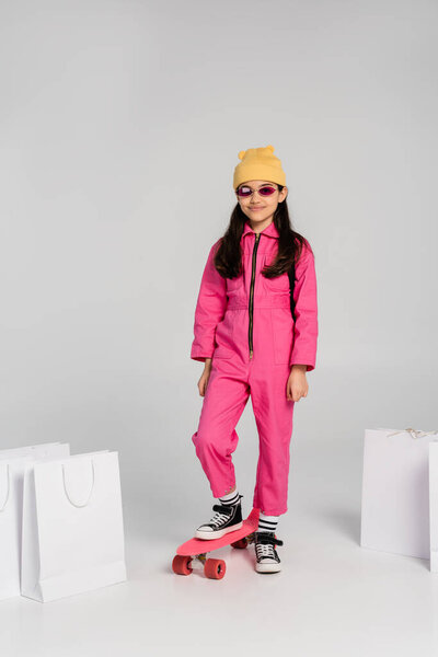 счастливая девушка в шапочке и стильных солнцезащитных очках верхом на доске, сумки для покупок на сером фоне