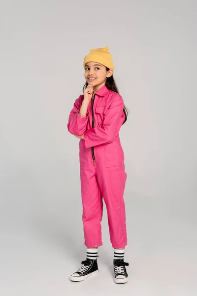 stock image happy kid in beanie hat and pink outfit standing and looking away, thinking on grey backdrop