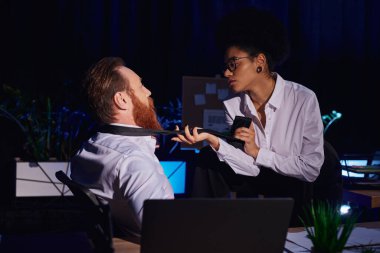 Seksi Afrikalı Amerikalı kadın sakallı iş adamının kravatını dizüstü bilgisayarın yanına çekiyor, gece ofisinde yakınlık kuruyor.