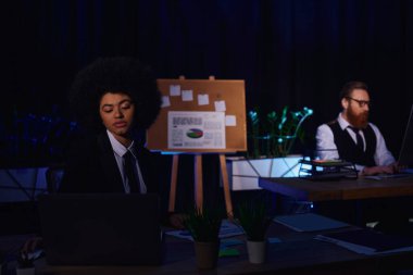 Çekici Afro-Amerikan iş kadını geceleri ofisin arka planında iş arkadaşının yanında çalışıyor.
