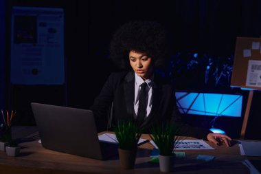 african american businesswoman in formal wear working on laptop in dark office, night shift clipart