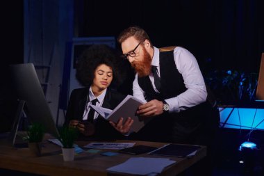 Sakallı iş adamı, gece ofisindeki bilgisayarın yanında Afro-Amerikan sekreterine defterini gösteriyor.