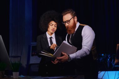 Afrikalı Amerikalı iş kadını, gece ofiste sakallı meslektaşının yanındaki belgeleri işaret ediyor.