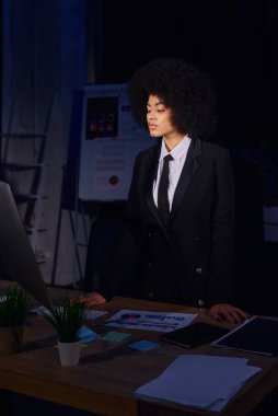 Genç Afrikalı Amerikalı iş kadını gece ofisindeki belgelerin yanındaki bilgisayar ekranına bakıyor.