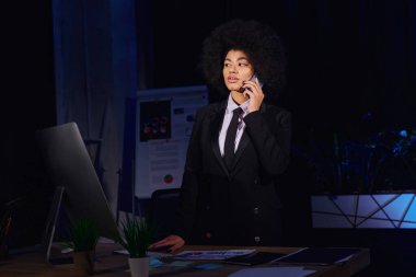 Afrika kökenli Amerikalı iş kadını gece ofiste bilgisayar monitörünün yanında akıllı telefondan konuşuyor.