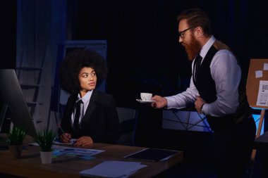 bearded businessman holding coffee near pleased african american secretary, flirting in night office clipart