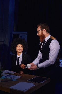 Sakallı iş adamı elinde kahve tutuyor. Afro-Amerikan sekreteri, gece ofisinde flört ediyor.