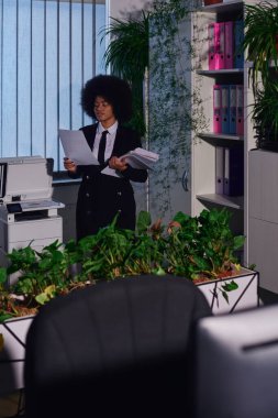 african american businesswoman with documents near copier and decorative plants at night in office clipart
