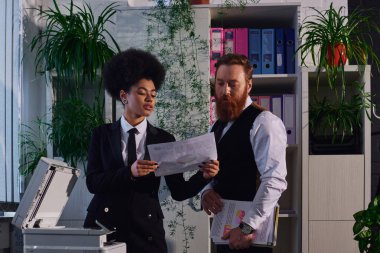 Genç Afro-Amerikan sekreteri ofisteki fotokopi makinesinin yanındaki sakallı iş adamına belge gösteriyor.