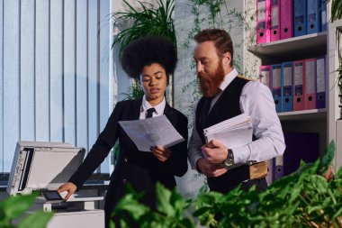 african american businesswoman with bearded colleague working with documents near copier in office clipart