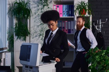 bearded businessman looking at attractive african american secretary near copier at night in office clipart