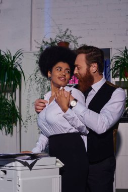 Sakallı iş adamı, fotokopi makinesinin yanında gülümseyen Afrikalı Amerikalı kadını kucaklıyor.