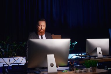 Ciddi sakallı iş adamı ofiste geç saatlere kadar çalışırken bilgisayar ekranına bakıyor.