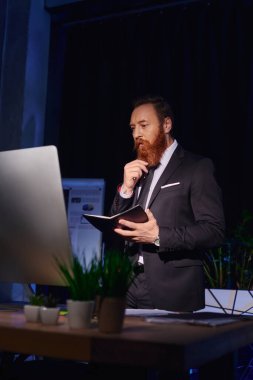 Düşünceli sakallı iş adamı. Not defteriyle bilgisayar ekranına bakıyor ve gece ofisinde çalışıyor.