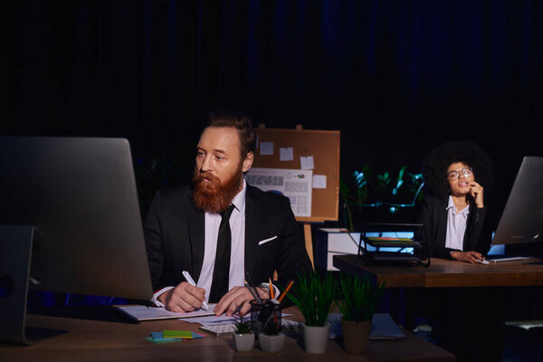 seductive african american woman looking at bearded businessman working at laptop at night in office