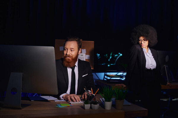 attractive african american woman near bearded businessman working on computer at night in office