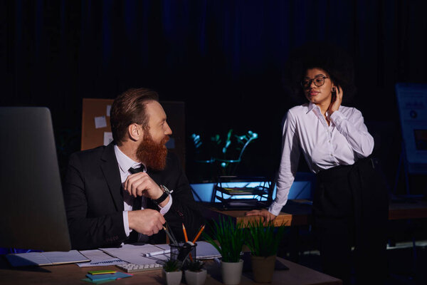 smiling bearded businessman looking at charming african american colleague in office at night
