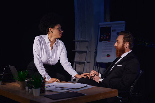 charming african american woman sitting on work desk and touching bearded businessman, seduction
