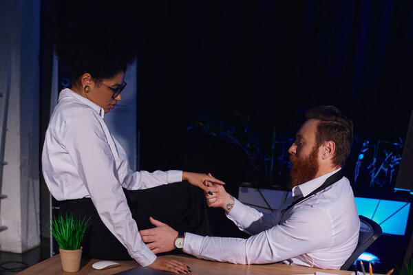 bearded businessman holding hand of african american woman sitting on desk, romance in night office