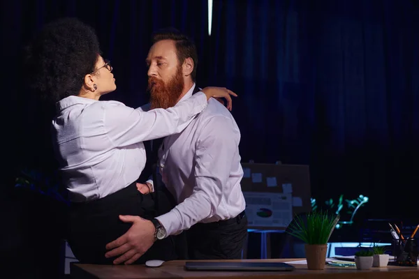 stock image bearded businessman embracing hot african american woman sitting on desk, seduction in night office