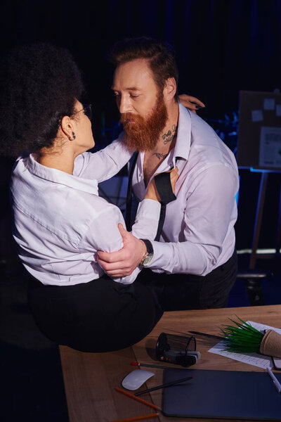 bearded tattooed man seducing african american woman near mess on work desk in office, love affair