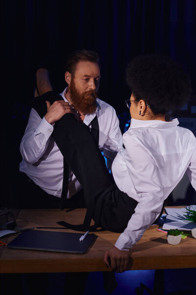 interracial couple flirting in office, businessman hugging leg of african american woman on desk