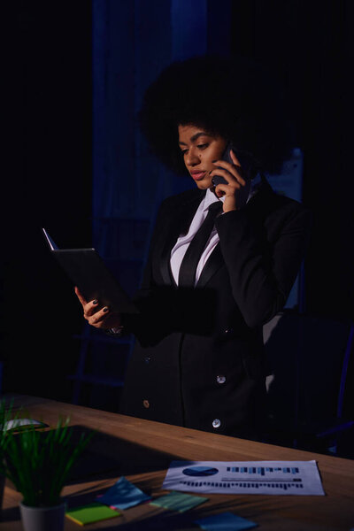 african american businesswoman with notebook talking on smartphone while working in office at night