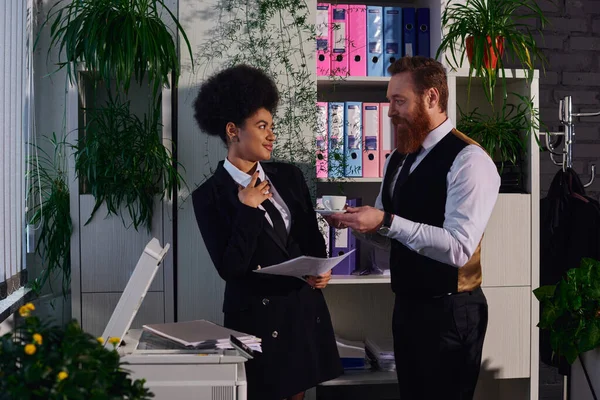 Stock image bearded businessman holding coffee cup and flirting with african american secretary in office