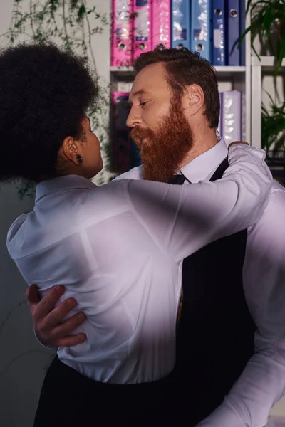 bearded businessman hugging young african american secretary, night romance in office