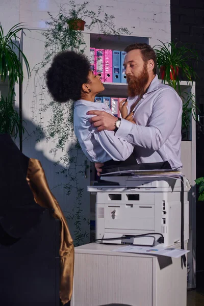 secret love in office, bearded businessman and african american woman embracing near copier at night