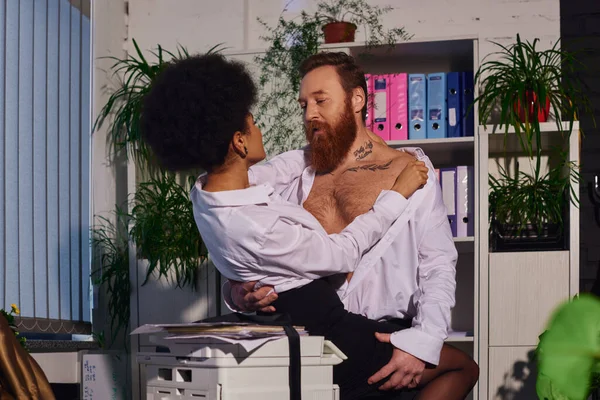 bearded tattooed businessman hugging young african american woman near copier, passion in office