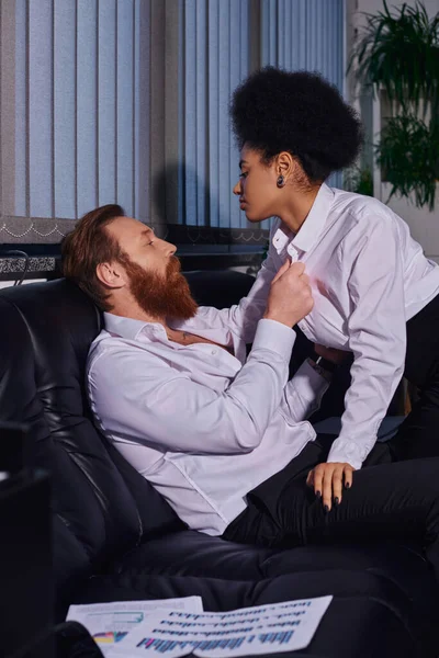 side view of charming african american woman seducing bearded businessman on couch in night office