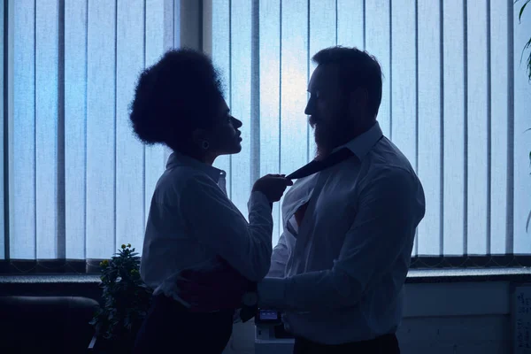 dark silhouette of african american woman pulling tie of colleague and seducing him in office