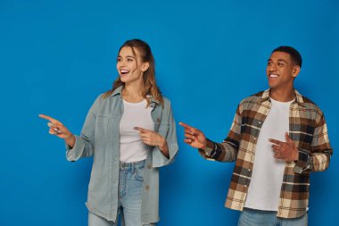 happy woman with open mouth and african american man pointing away with fingers on blue background clipart