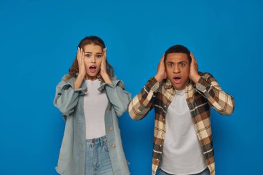 shocked interracial couple with open mouth looking at camera on blue background, emotional reaction clipart