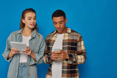 Mavi arka planda dururken akıllı telefon ve dijital tablet kullanan mutlu ırklar arası çift