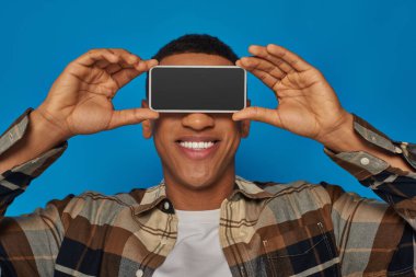 Mutlu Afro-Amerikalı adam yüzünü gizliyor mavi arka planda akıllı telefonuyla, belirsiz yüzüyle.