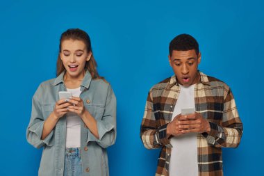 Heyecanlı Afro-Amerikan erkeği ve şaşkın genç kadın mavi arka planda akıllı telefonlarda sohbet ediyorlar.