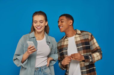 Mutlu Afrikalı Amerikalı adam, bayan arkadaşının akıllı telefonuna bakıyor ve mavi arka planda gülümsüyor.