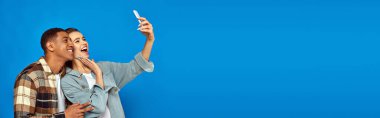 cheerful interracial couple taking selfie while looking at smartphone on blue backdrop, banner clipart
