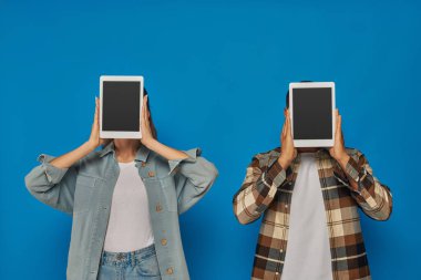 Irklar arası kadın ve erkek, yüzlerini gizleyen dijital tabletlerle mavi arka planda, boş dokunmatik ekranda