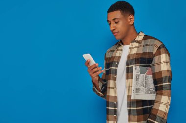 Ekose gömlekli Afro-Amerikan adam akıllı telefon kullanıyor ve mavi arka planda gazete tutuyor.