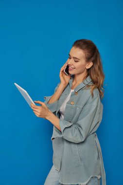 Akıllı telefondan konuşan ve mavi arka planda dijital tablet kullanan mutlu genç bir kadın.