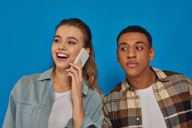Afro-Amerikalı adam mavi arka planda akıllı telefondan konuşan mutlu kadının konuşmasını dinliyor.