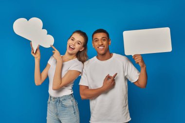 amazed interracial couple pointing at blank placards on blue backdrop, thought and speech bubbles clipart