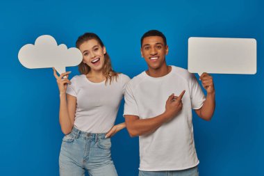 excited interracial couple pointing at blank placards on blue backdrop, thought and speech bubbles clipart